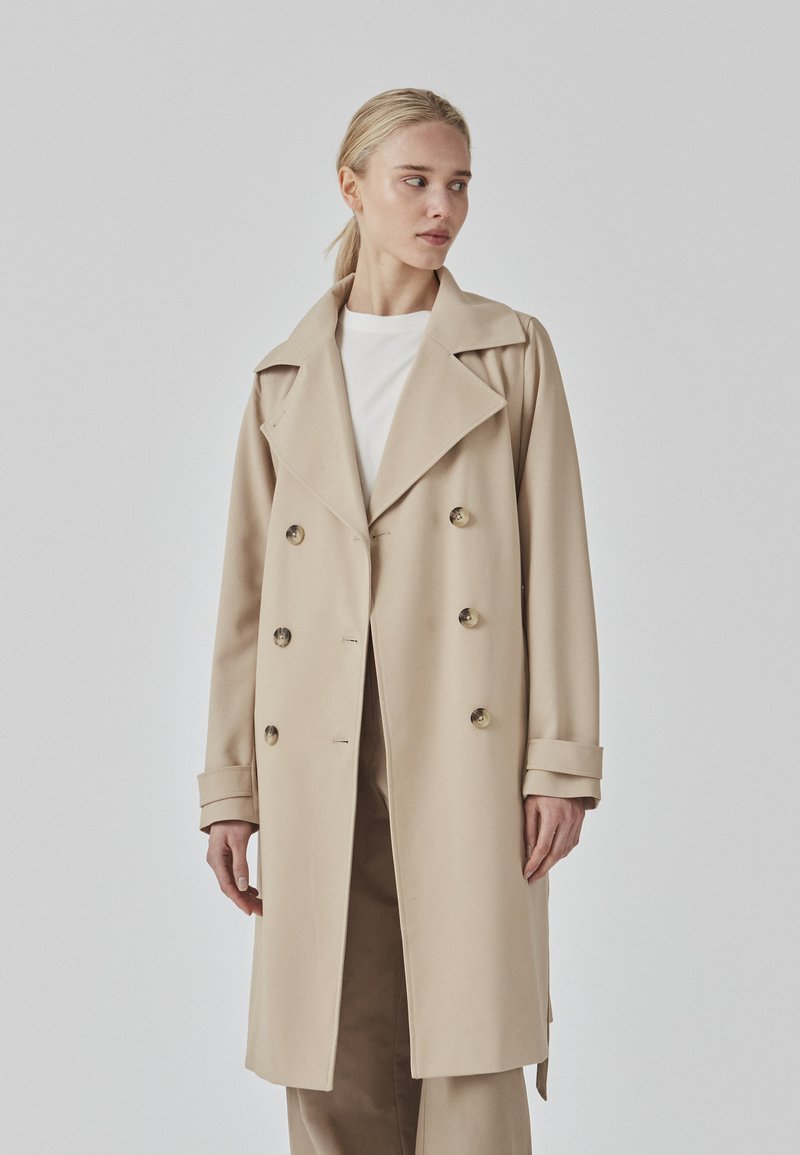 Beige trench coat with a double-breasted design, large lapels, and button accents. Features wide cuffs and straight hemline. Smooth texture.