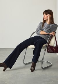 Pull gris en tricot, pantalon large bleu marine foncé, bottines à talons marron à motif et sac à main bordeaux, assise sur une chaise chromée.