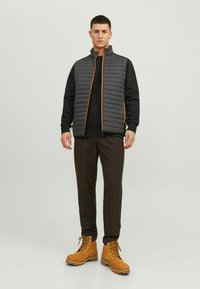 Grau gestepptes Weste mit einem orangefarbenen Reißverschluss, kombiniert mit einem schwarzen Pullover und dunkelbraunen Hosen. Braune Schnürstiefel runden das Outfit ab.