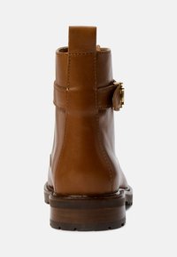 Bota de tornozelo em couro castanho com um acabamento suave, apresentando um destaque em fivela e uma sola de madeira robusta. Salto alto com uma aba na parte traseira.