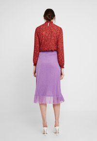 Červená květinová halenka s vysokým límcem a dlouhými rukávy, doplněná levandulovou třpytivou sukní s průsvitným volánovým lemem, nošená s bílými podpatky.