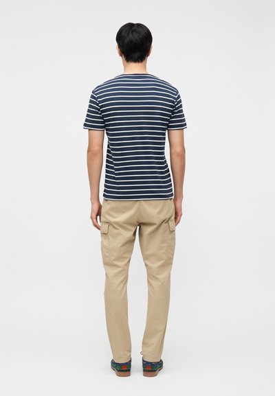 Άνδρας με την πλάτη στραμμένη, φορά μπλε σκούρη και λευκή ριγέ μπλούζα, μπεζ παντελόνι cargo και πράσινα παπούτσια μπροστά σε απλό ανοιχτό φόντο.