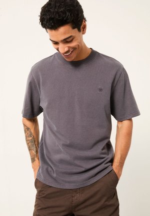 Mann mit tätowiertem Unterarm, trägt ein graues T-Shirt mit Rundhalsausschnitt und kurzen Ärmeln sowie braune Hose, lächelt und schaut nach unten, die Hände in den Taschen.