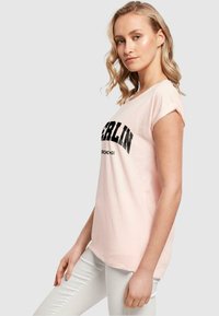 Lyserød t-shirt med korte, rullede ærmer, som har et iøjnefaldende sort "BERLIN" grafik og en blød stofstruktur.