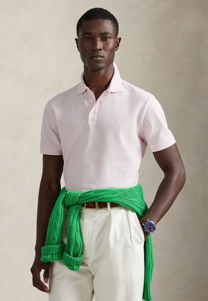 Īsroku krekliņš ar svītrām gaiši rozā krāsā, kopā ar zaļu pīta adījuma džemperi, sasietu viduklī, un baltām biksēm.