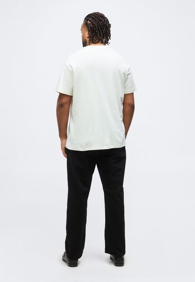 Άνδρας με πλεξούδες στέκεται με την πλάτη γυρισμένη, φοράει απλό λευκό κοντομάνικο πουκάμισο, μαύρο παντελόνι και μαύρα παπούτσια σε λευκό φόντο.