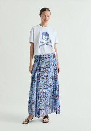 Femme portant un t-shirt blanc avec un imprimé tête de mort et os croisés bleus et une longue jupe bleue à motifs, debout devant un fond uni.