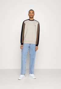 Sudadera en diseño bicolor beige, marrón y negro; cuello redondo, mangas largas. Combinada con vaqueros rectos de tiro medio en azul claro y zapatillas blancas.