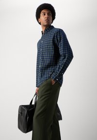 Karierte blaue und grüne Hemd mit Button-Down-Kragen, Fronttasche, getragen mit grünen Hosen, hält eine schwarze Lederhandtasche.