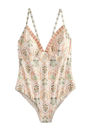 Frau trägt geblümten Badeanzug und weißes Kopftuch und steht an einem Holztisch mit Tellern und Glas im Freien im Sonnenlicht.