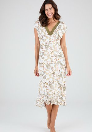 Femme souriante, portant une robe midi blanche à imprimé floral vert et un col en V en dentelle vert olive, debout pieds nus sur un fond uni.