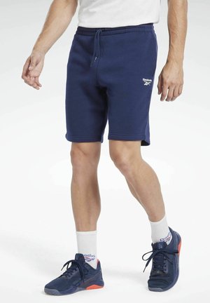 Mann trägt marineblaue Reebok-Shorts, weiße Socken und marineblaue Laufschuhe mit orangefarbenen Akzenten und steht vor weißem Hintergrund.