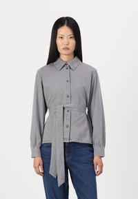 Grå denimskjorta med långa ärmar, knäppning fram, spetsig krage och ett knytband runt midjan. Har en mjuk textur.