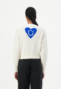 Pull en maille blanche avec un motif de cœur bleu comportant les lettres "HGO" et un smiley. Poignets et ourlet côtelés. Coupe décontractée.