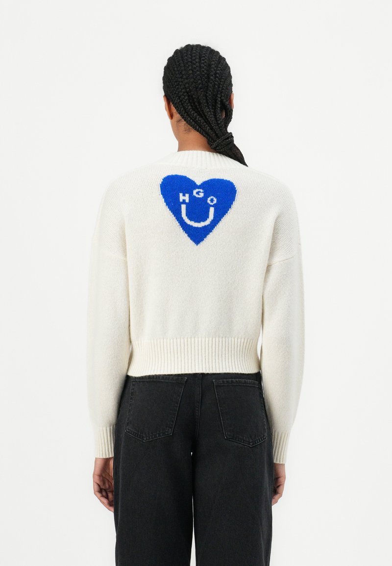 Pull en maille blanche avec un motif de cœur bleu comportant les lettres "HGO" et un smiley. Poignets et ourlet côtelés. Coupe décontractée.