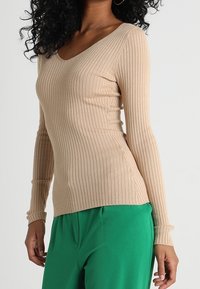 Beige žebrovaný top s dlouhým rukávem a výstřihem do V, v kombinaci se zelenými kalhotami. Materiál je texturovaný a těsný, vytvářející štíhlou siluetu.