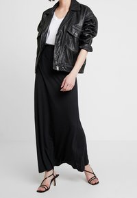Chaqueta oversized de cuero negra con cremallera y detalles de bolsillos visibles, combinada con una falda longa negra y tacones negros con tiras. Camiseta blanca debajo.