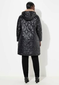 Gequiltede zwarte pufferjas met een tan nepbont voering in de capuchon, lange mouwen en een knielange snit, gecombineerd met zwarte broek en hakken.