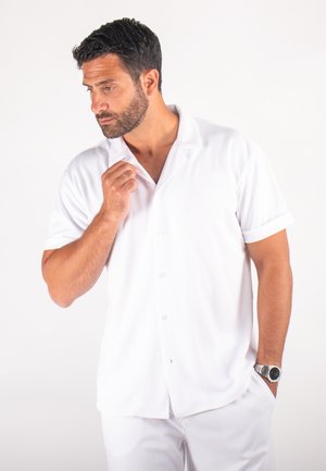Chemise blanche à manches courtes avec col, en tissu texturé et manches retroussées, portée sur un pantalon blanc coordonné.