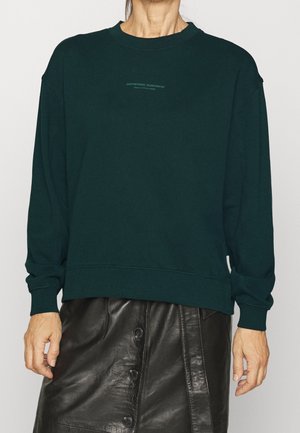 Person iført en mørkegrøn crewneck-trøje med lille tekst og en sort lædernederdel med knapper.