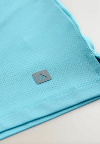 Camisa azul claro de manga corta hecha de un tejido transpirable con textura de malla, que presenta un logo rectangular gris en el lado inferior derecho.