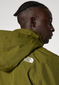 Olivově zelená bunda s kapucí z texturované látky. Na zadním rameni je v bílé barvě vyobrazeno logo The North Face.