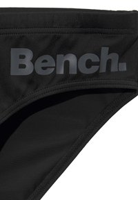 Bas de bikini noir avec une finition textile lisse. Présente un logo "Bench." gris bien visible sur la ceinture. Design simple avec un minimum de couture.