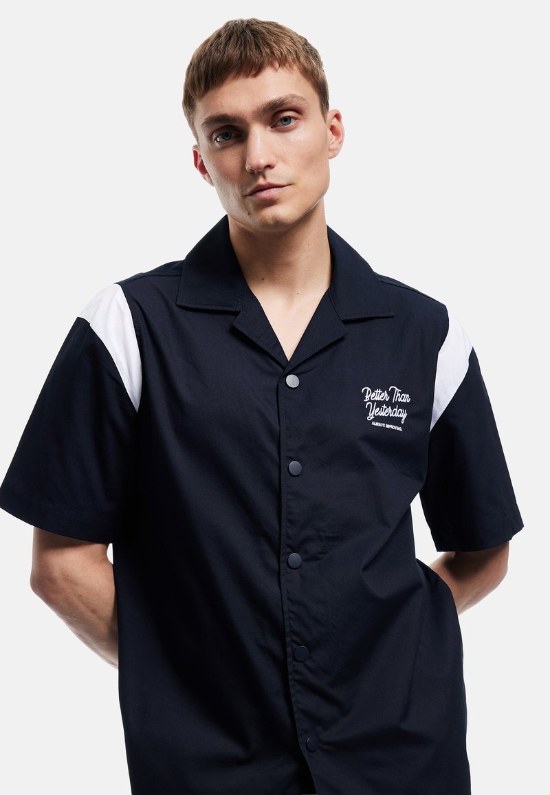 Jeune homme aux cheveux courts châtain clair portant une chemise noire à manches courtes avec des accents blancs sur les épaules et le texte « Better Than Yesterday ».
