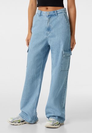 Lyseblå vidbenede denim cargobukser båret med hvide og grønne sneakers og en sort cropped top, vist på en stående person.