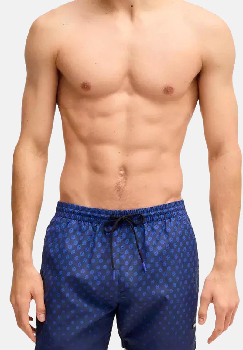 Homme torse nu avec des abdos définis portant un short de bain bleu avec un petit motif circulaire et un cordon noir, debout devant un fond uni.