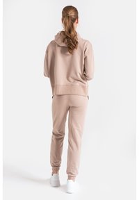 Jasno beżowy sweatshirt z kapturem i dopasowanymi dresami, wykonany z miękkiego materiału. Posiada elastyczne mankiety i luźny krój. Nosi się z białymi sneakersami.