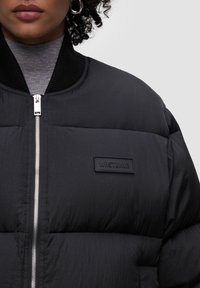 Czarna kurtka puchowa o teksturowanym wykończeniu, z zapięciem na suwak z przodu, prążkowanym kołnierzem i gumową naszywką z logo na piersi.