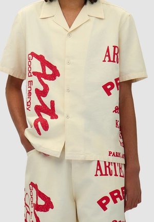 Chemise à manches courtes, crème avec du texte et des motifs en paillettes rouges. Elle possède un col classique et une fermeture à boutons, fabriquée en tissu léger.