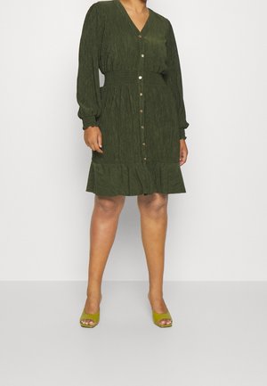 Kobieta ubrana w teksturowaną, ciemnozieloną sukienkę z długim rękawem z przodu zapinaną na guziki i z falbanką u dołu, zestawioną z zielonymi, otwartymi na palce szpilkami.