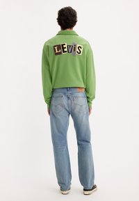 Pullover in pile verde con una toppa colorata "Levi's" sul retro. Indossato con jeans denim azzurri e sneakers nere. Vestibilità comoda.