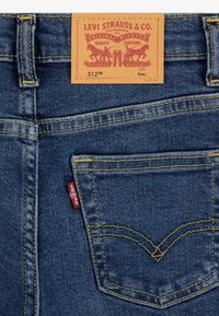 Siniset farkut keltaisella ompeleella, joissa on takatasku erottuvalla V-muotoisella kuviolla ja kiinnitetty ruskea merkkilappu.