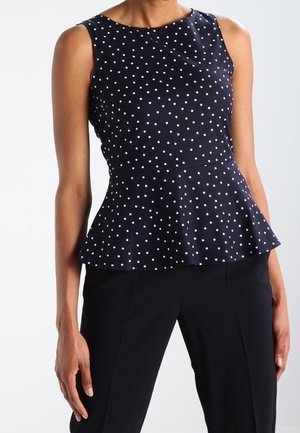 Granatowy top bez rękawów z baskinką i białymi groszkami, noszony z czarnymi spodniami na stojącej osobie na jednolitym tle.