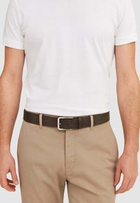 Cintura marrone intrecciata con fibbia rettangolare in argento, indossata sopra pantaloni kaki e una maglietta bianca, che mette in risalto la trama e i dettagli del design.