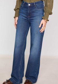 Jean en denim bleu foncé, taille haute, jambes larges. Dispose de poches avant, d'un système de fermeture à bouton et d'un léger dégradé sur le tissu.