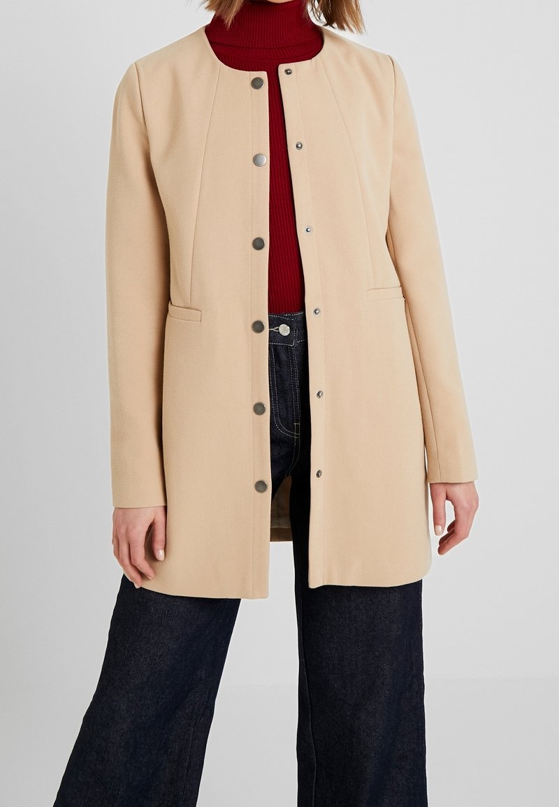 Abrigo beige con un corte entallado, cuello redondo, cierres de broche y bolsillos laterales. El material parece texturizado y suave. Pantalones de mezclilla oscuros debajo.