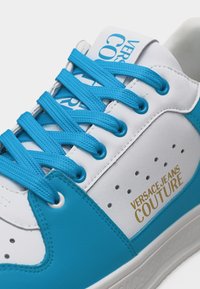 Άσπρο και μπλε αθλητικό παπούτσι με μπλε κορδόνια, λογότυπο "Versace Jeans Couture" σε χρυσό στο πλαϊνό πάνελ και μπλε λογότυπο στη γλώσσα.