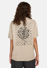 Beige cotton t-shirt with a black graphic design featuring vines over a diamond-shaped grid pattern on the back. Short sleeves and relaxed fit.