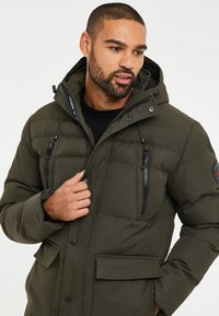 Mørkegrøn pufferjakke med hætte, lynlåslommer og trykknaplukninger. Stoffet er glat med en mat finish og subtile syninger.