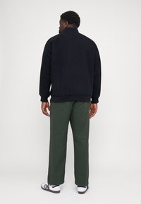 Casaco de forro polar preto com colarinho alto, usado sobre calças verdes-azeitona. O casaco tem punhos canelados e um corte descontraído. Ténis brancos com riscas pretas.