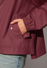 Vínovočervená nepremokavá bunda s elastickými manžetami, bočným vreckom a hladkou textúrou, zobrazená na neutrálnom pozadí.