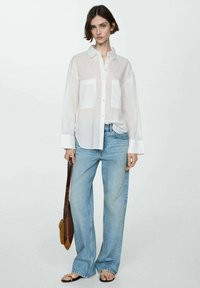 Camicia bianca trasparente con bottoni e due tasche frontali, abbinata a jeans ampi blu chiaro e sandali piatti. Borsa a tracolla marrone come accessorio.