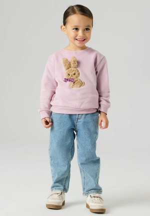 Jasnofioletowa bluza z teksturowaną aplikacją zajączka o brązowym futerku i fioletowej kokardce, zestawiona z niebieskimi dżinsami i białymi butami.