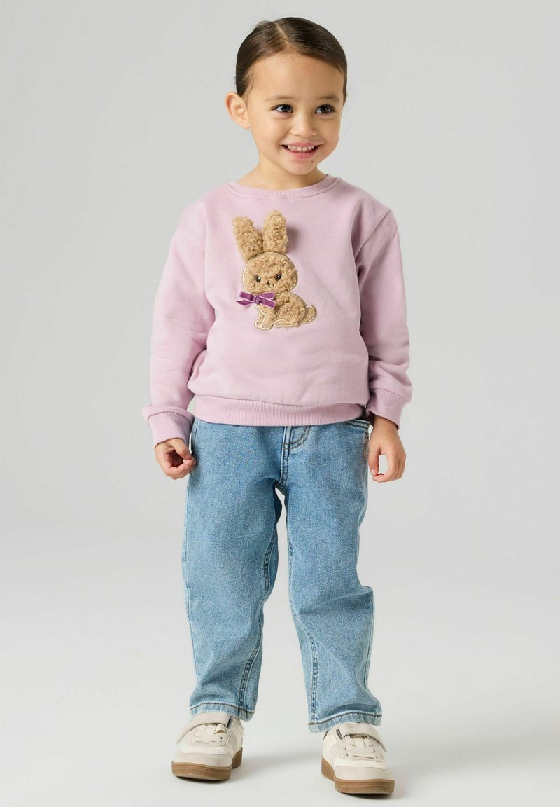 Helelilla dressipluus, millel on tekstuuriga jänkuapplikaatsioon pruunide karvade ja lillaka vööga, kombineeritud siniste teksade ja valgete kingadega.