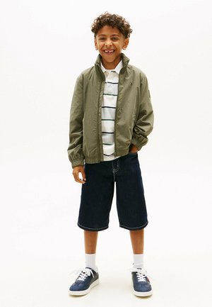 Lächelnder Junge mit lockigem Haar, der eine olivgrüne Jacke, ein weiß gestreiftes Polohemd, dunkle knielange Shorts, weiße Socken und marineblaue Turnschuhe trägt.