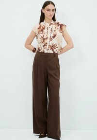 Bloemige blouse in crème en bruin met korte mouwen, gecombineerd met hoog in de taille zittende, wijde bruine broek en bruine puntige schoenen.